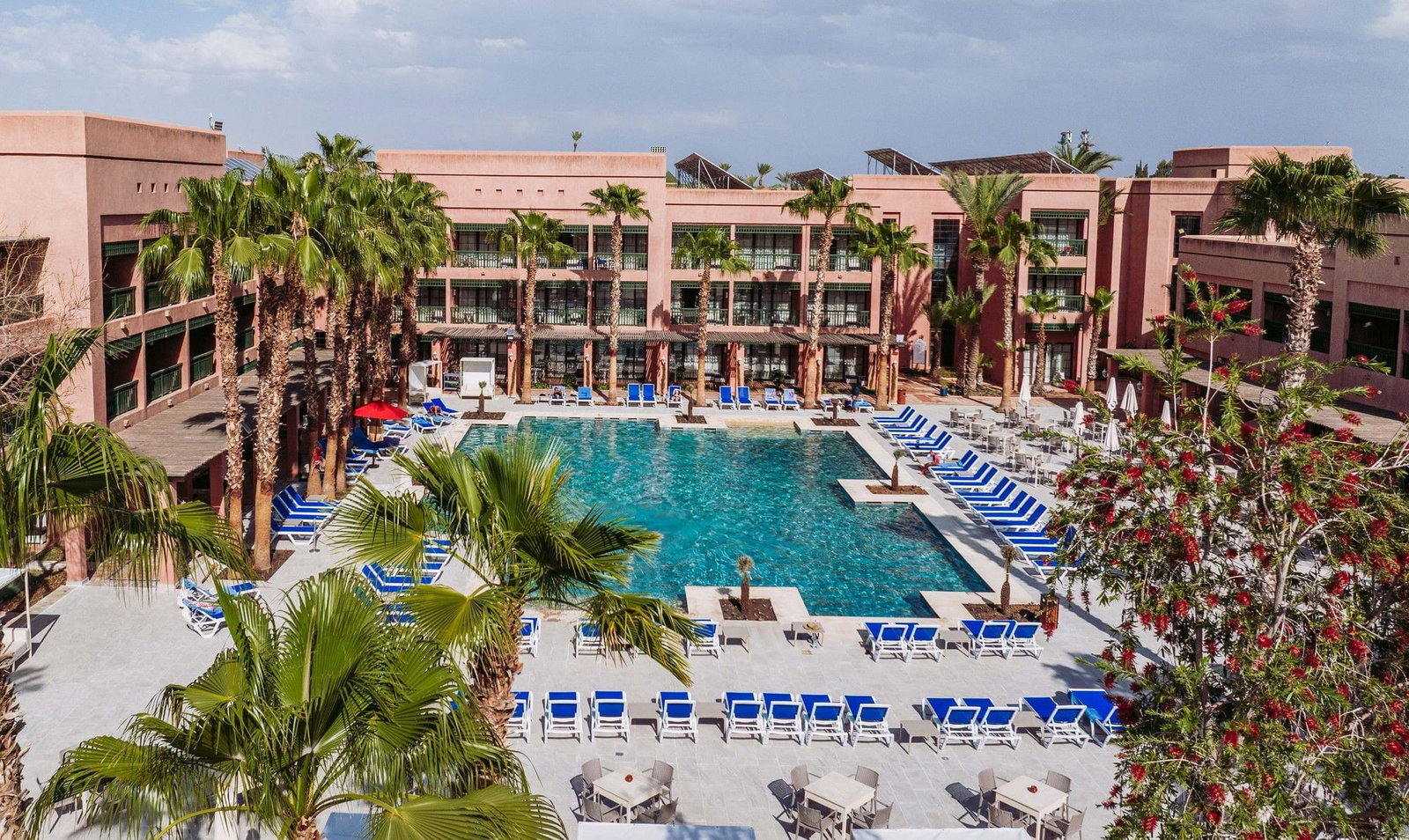 Aerial view of a sunlit hotel courtyard with a large rectangular swimming pool, rows of blue sunbeds and palm trees