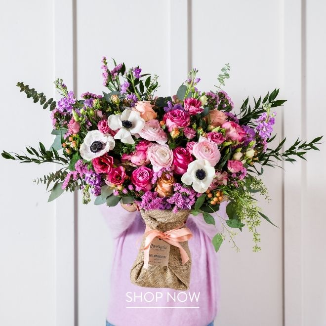 A person holding a tall burlap-wrapped bouquet of pink roses, ranunculus, white anemones, purple stock, and eucalyptus, tied with a peach ribbon, against a white paneled wall