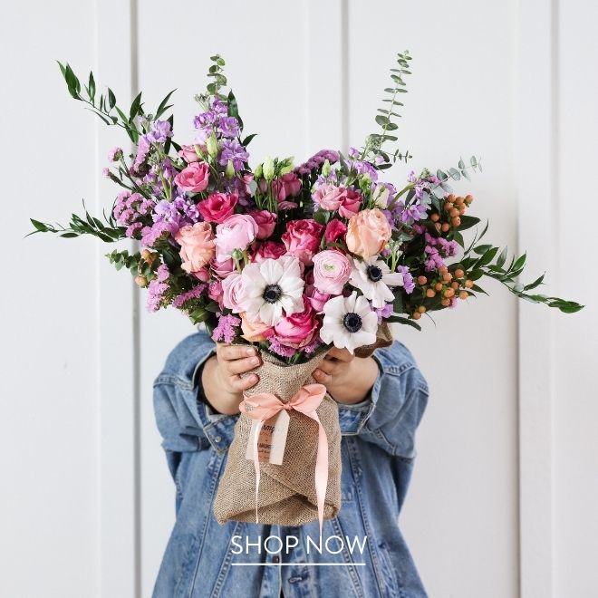 A person in a denim jacket holding a large burlap-wrapped bouquet up in front of their face, filled with pink roses, ranunculus, white anemones, purple stock, and eucalyptus, tied with a peach ribbon