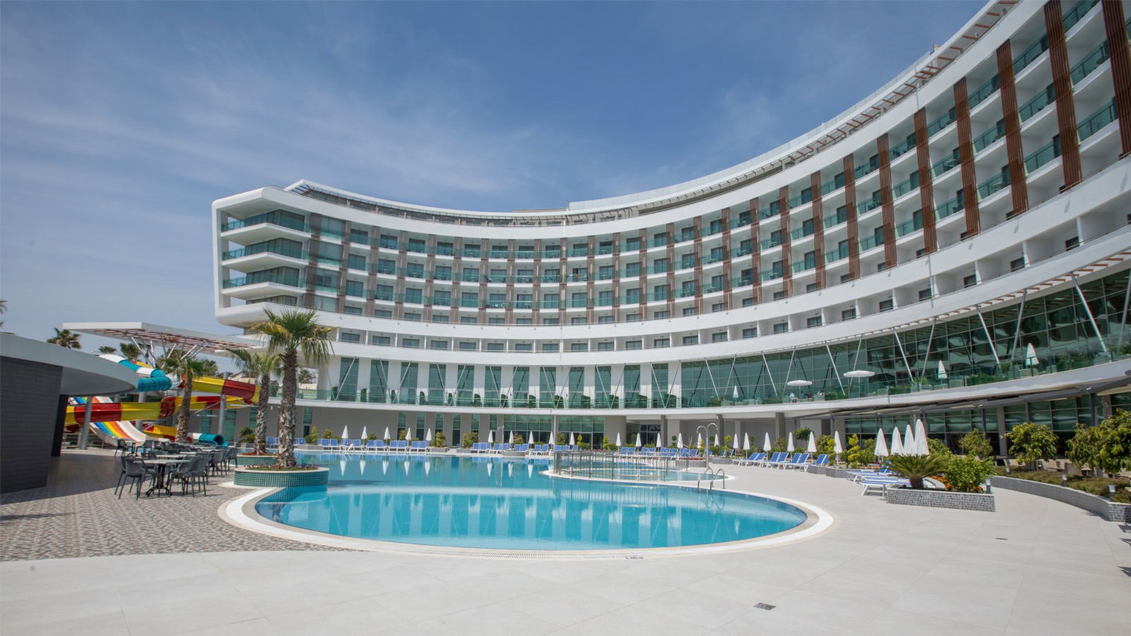 Curved modern hotel with a circular outdoor pool, sunloungers, palm trees and colourful water slides under a blue sky