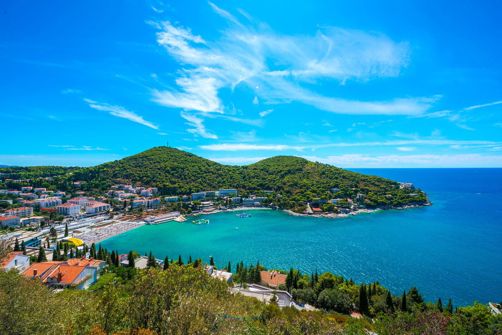 Aerial view of a Mediterranean seaside resort with turquoise bay, sandy beach, green hills and coastal villas under a bright blue sky