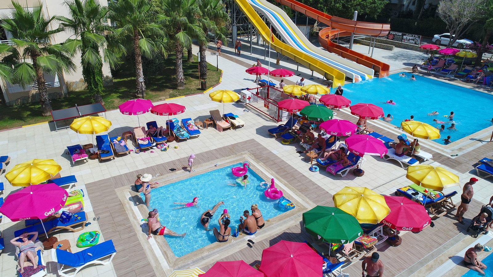 Aerial view of busy hotel waterpark with colourful parasols, sunloungers, two pools and tall water slides, families enjoying summer
