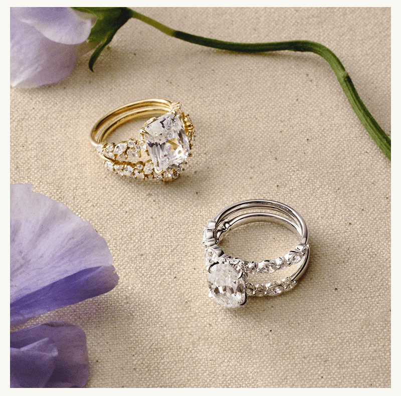 Diamond ring stacks, in yellow gold with radiant stone & white gold with oval stone, arranged next to flowers.