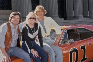 American actor, and politician Ben Jones, wearing a brown leather waistcoat over a blue shirt with rolled-up sleeves and jeans, beside a woman who wears a white-fringed black top, jeans and sunglasses, and American actor and singer John Schneider, in an unbuttoned pale yellow shirt over a blue t-shirt with stonewashed jeans, all three sitting on the General Lee, United States, January 1981.