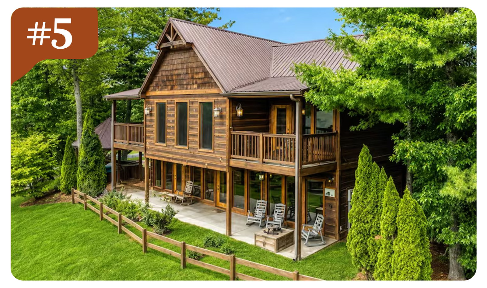 Rustic two-story woodland lodge with wraparound porches, rocking chairs, and lush greenery all around.