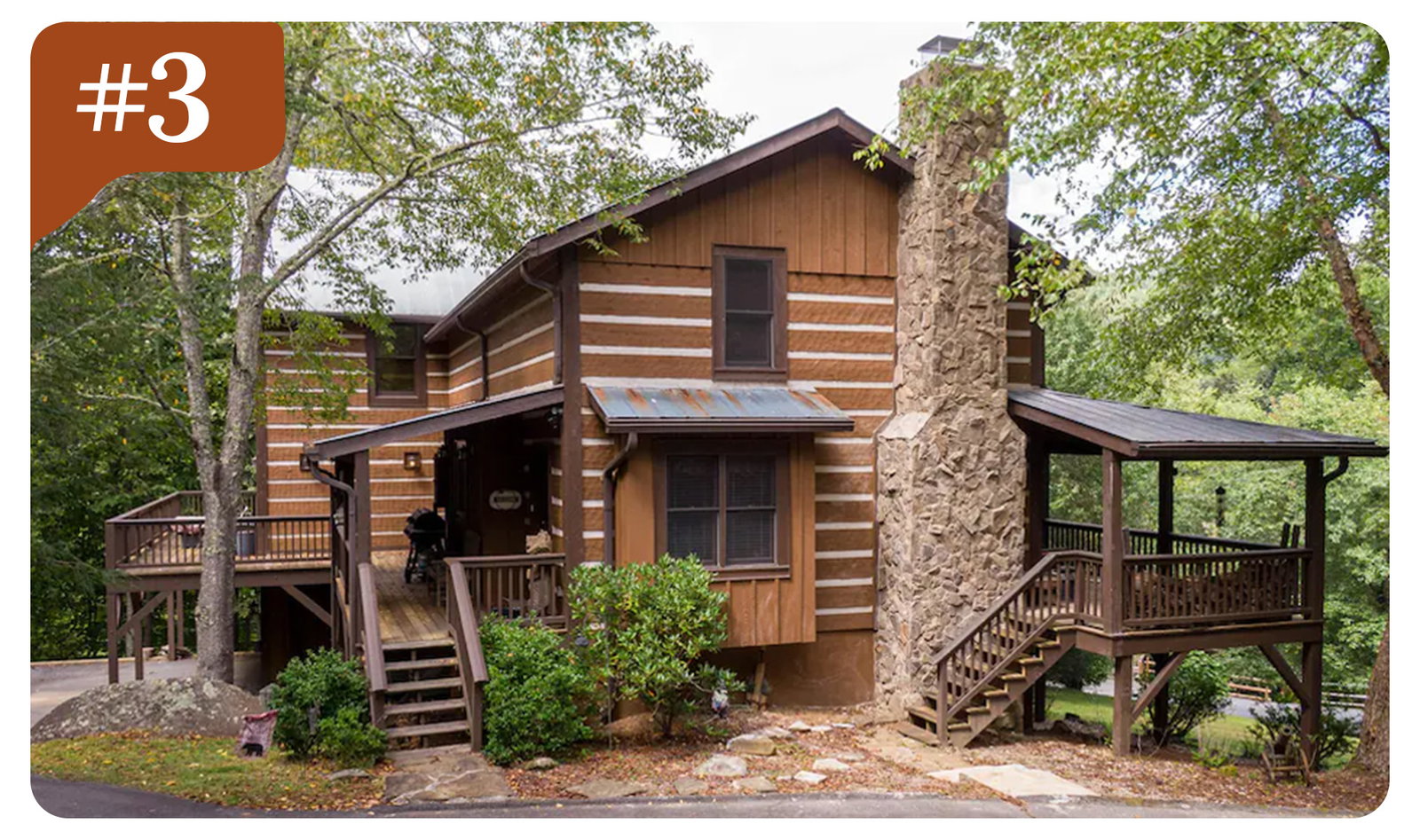 Rustic wood cabin with stone chimney, wraparound decks, and trees in a wooded setting.
