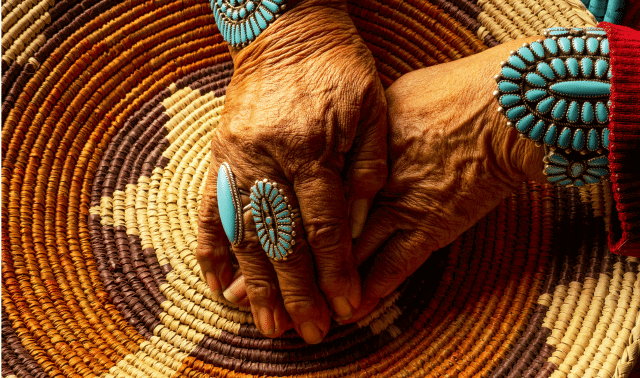 Elderly hands with turquoise jewelry rest on a woven basket with a geometric pattern in earth tones.