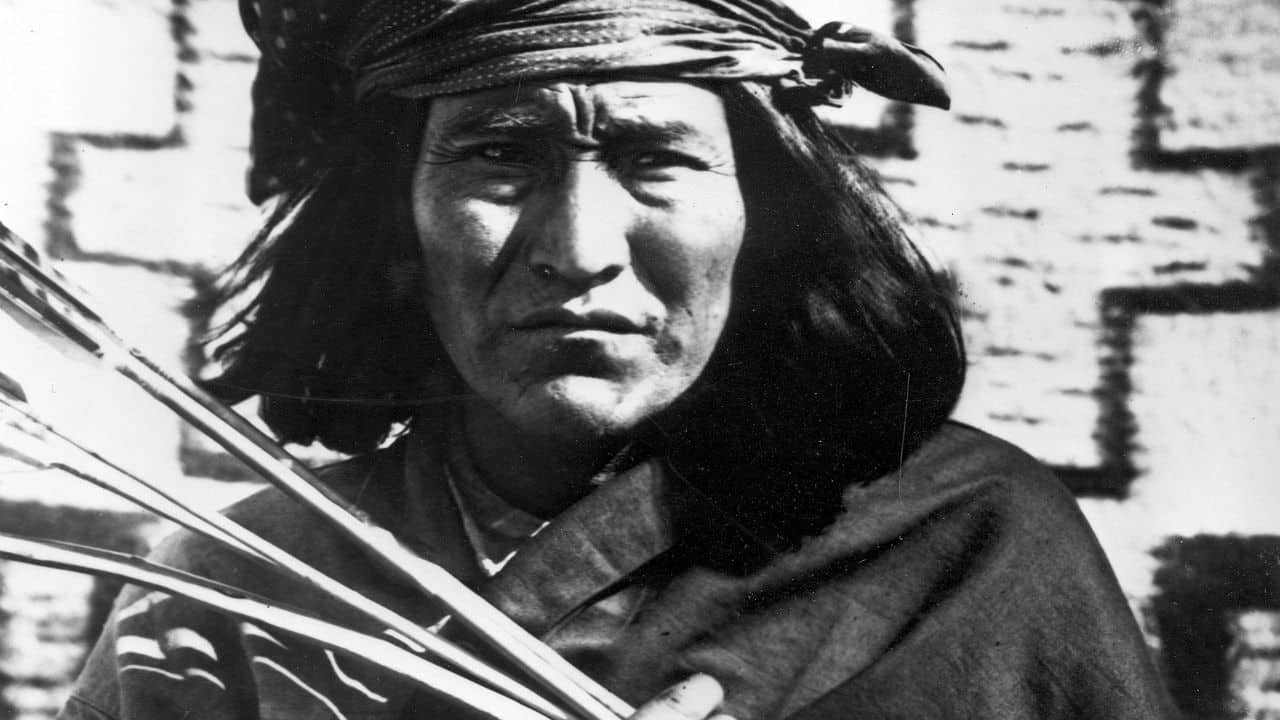 A person with long hair wearing a patterned headscarf and traditional clothing holds several arrows, standing in front of a geometric patterned background.