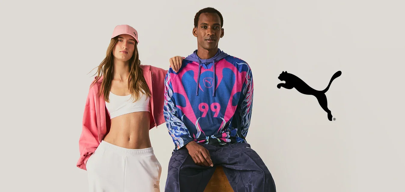Eine Frau in weißen Hosen, Crop-Top und pinker Jacke steht neben einem Mann, der auf einem Hocker sitzt und einen bunten Hoodie und dunkle Hosen trägt.