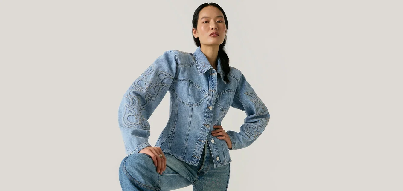 Frau mit langen dunklen Haaren, die eine hellblaue bestickte Jeansjacke und passende Jeans trägt, posiert mit einem erhobenen Knie vor einem einfarbigen Hintergrund.