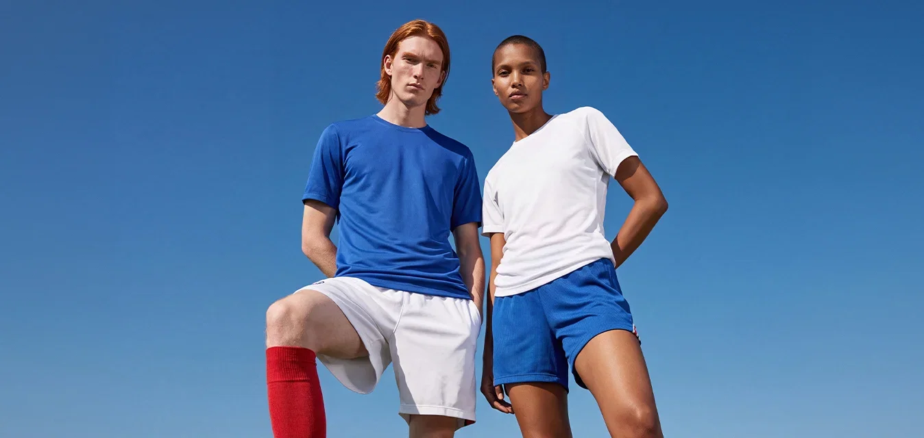 Zwei Fussballspieler stehen auf einem Grasfeld unter klarem Himmel. Der eine hat den Fuss auf dem Fussball und trägt rote Socken, der andere hat die Hände hinter dem Rücken.
