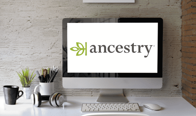 A desktop computer displays the Ancestry logo. The desk also holds various items including a plant, headphones, and a keyboard.