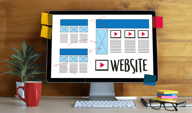 A computer monitor displays a website wireframe, with a plant, a red mug, and sticky notes around it on a wooden desk.