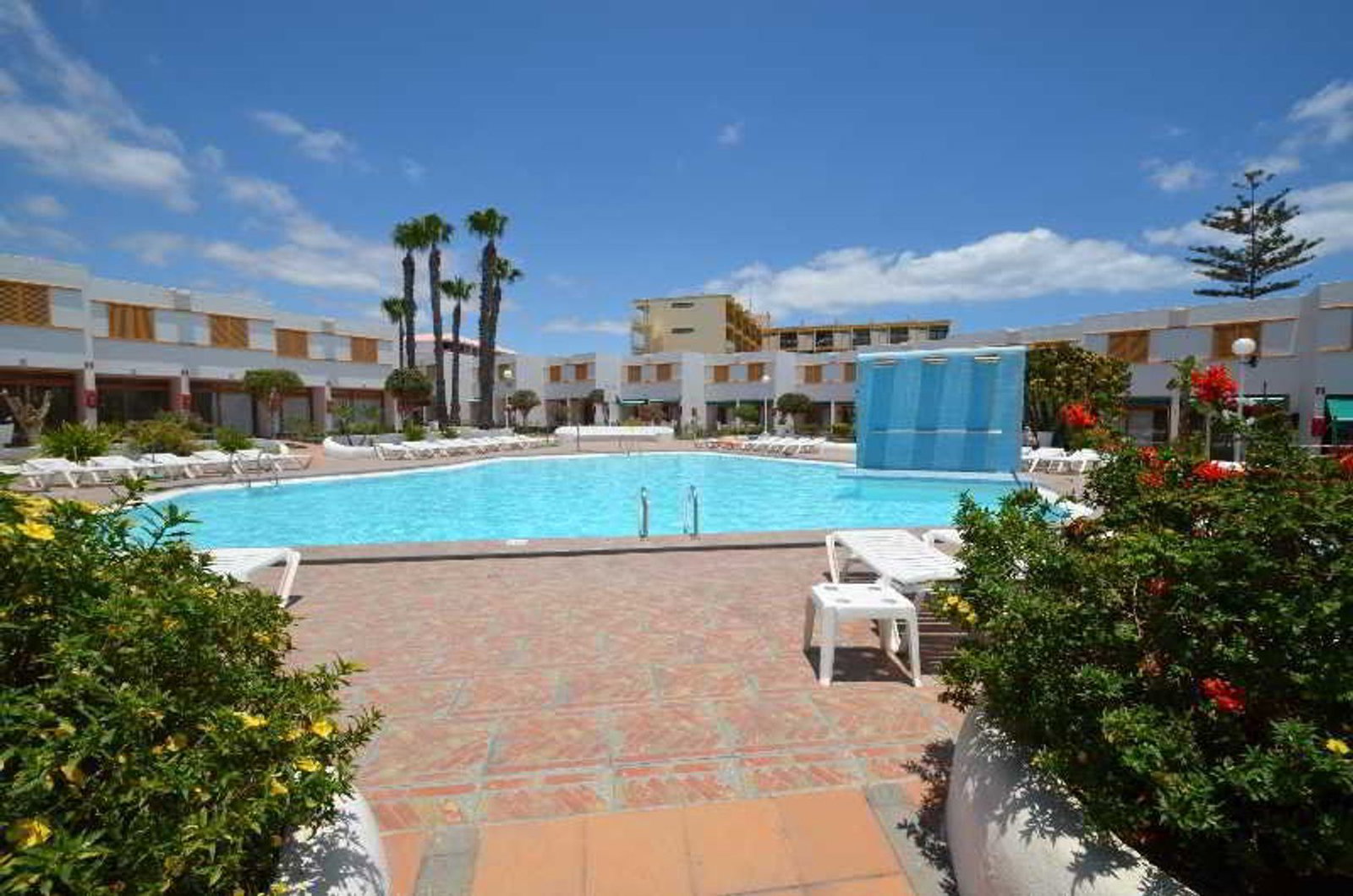 Outdoor hotel swimming pool with sun loungers, palm trees and white low-rise resort buildings under a bright blue sky