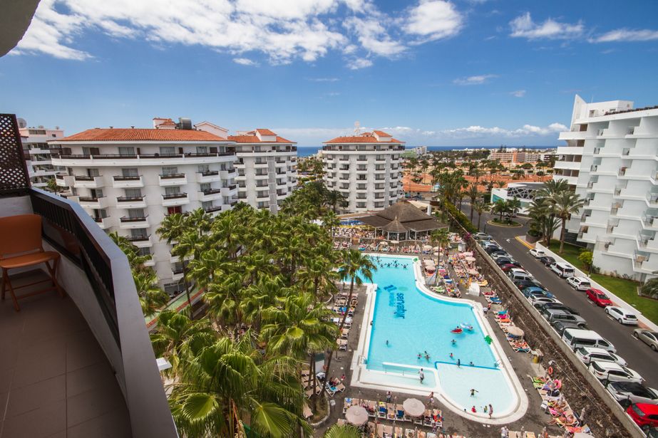 Balcony view of sunny resort with large pool, sunloungers, palm trees and white apartment blocks with sea in the distance