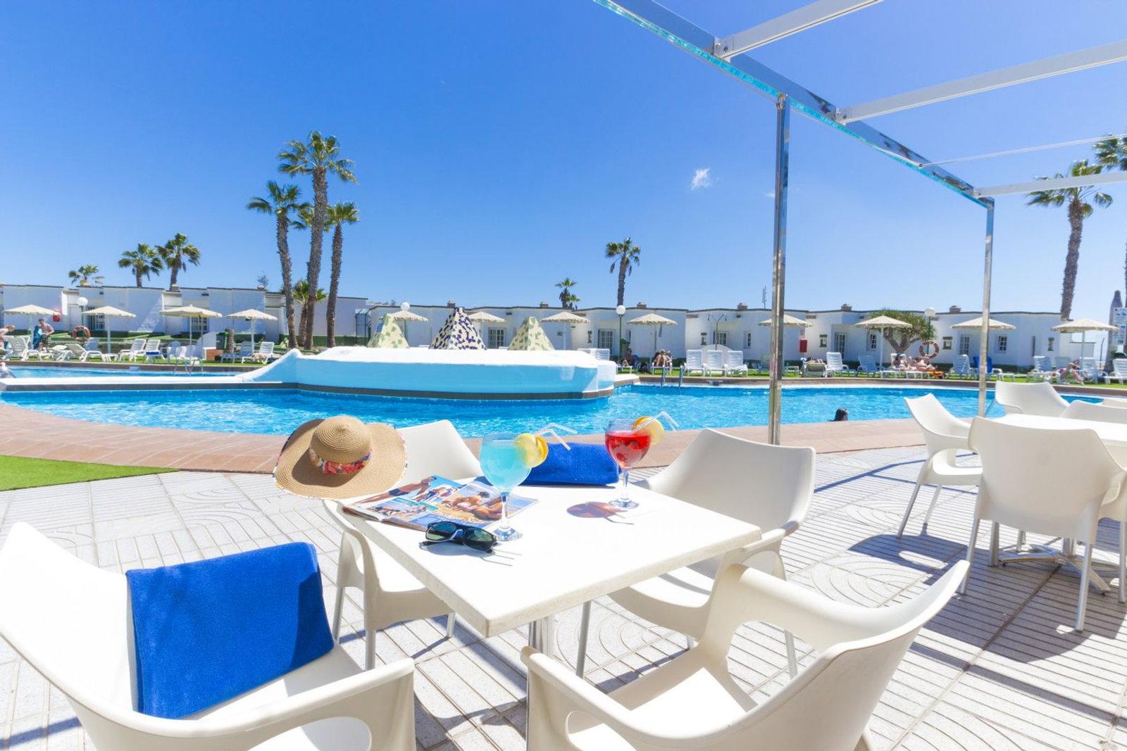 Sunlit resort poolside with white table, cocktails, sunhat, sunglasses, palm trees and loungers under blue sky