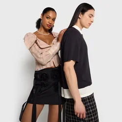 Zwei Modelle stehen Rücken an Rücken. Die Frau trägt ein hellrosa strukturiertes Oberteil und einen schwarzen Rock mit floralen Applikationen. Der Mann trägt ein schwarzes Hemd und karierte Hose.