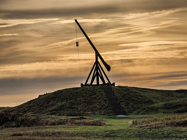 A dramatic sunset frames a large trebuchet perched on a grassy hill, creating a striking silhouette. 