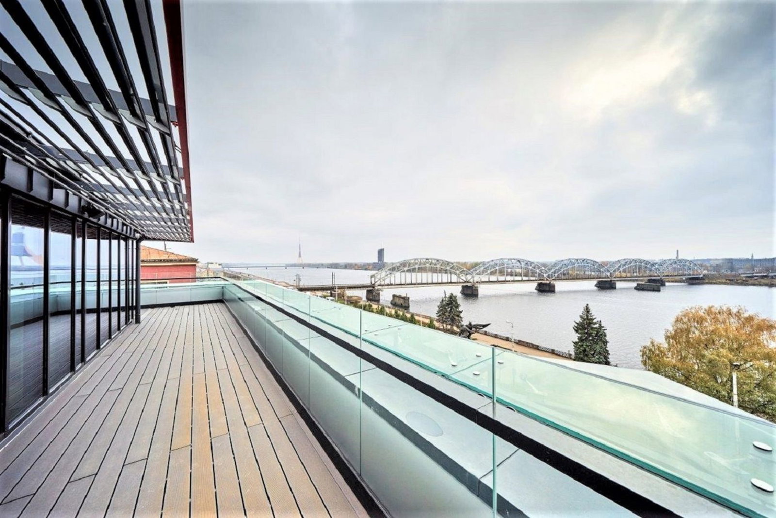 Spacious riverside rooftop terrace with glass balustrade, wooden decking and panoramic view of arched railway bridge and skyline.