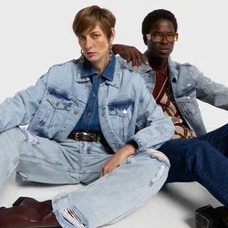 Twee modellen zittend op de vloer, gekleed in denim jassen en versleten jeans. Het eerste model heeft bruin haar, het tweede model draagt een bril en een patroon top.