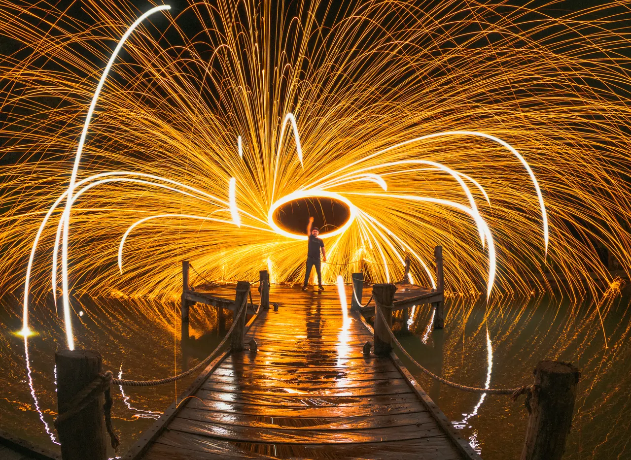 A person swinging burning steel wool to create a circular light display on a wooden pier at night.