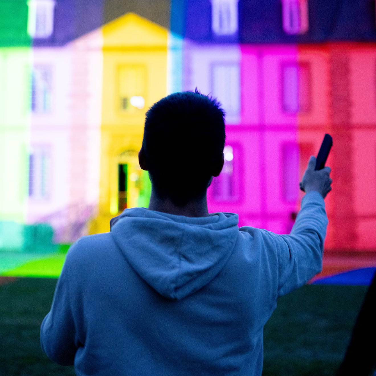 Philippe Dubost standing before a brightly illuminated building, holding up a device while testing colorful light projections during exhibition setup.