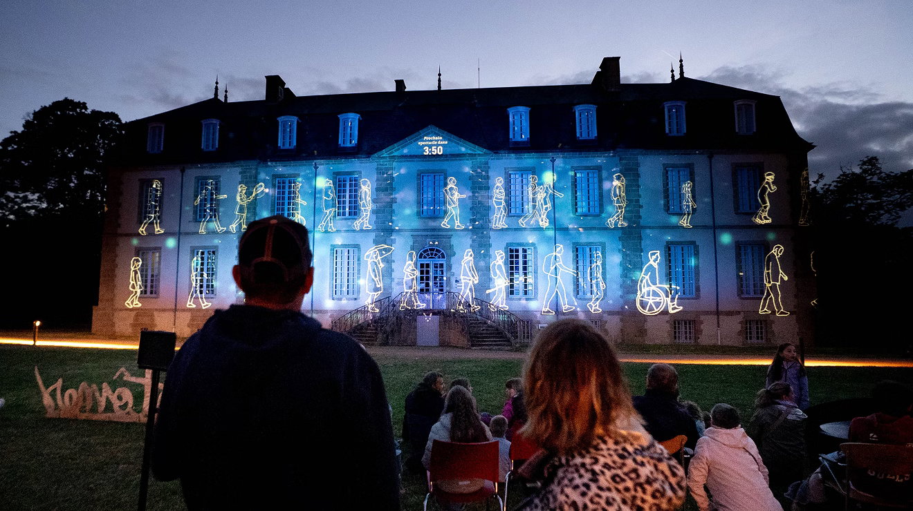 Audience members sit on a lawn at dusk watching a large projection mapped onto the front of a historic building. Animated outline figures appear to walk across the illuminated façade in shades of blue and white, creating an outdoor light show against the evening sky.