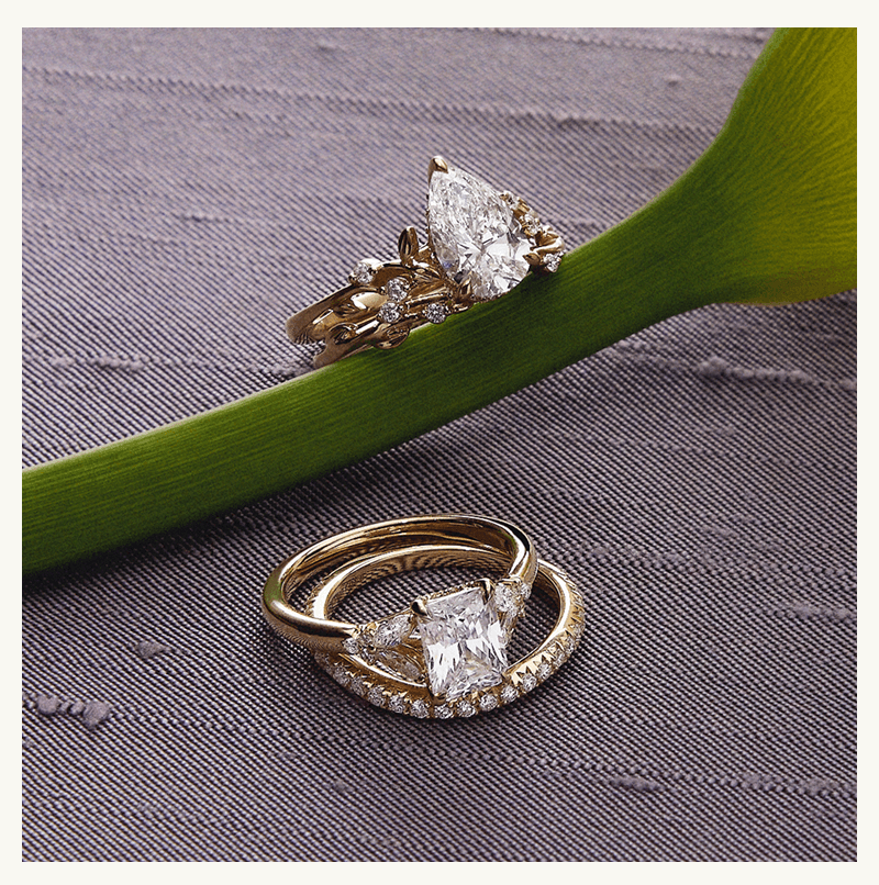 Diamond ring stacks, in yellow gold with radiant stone & white gold with oval stone, arranged next to flower.