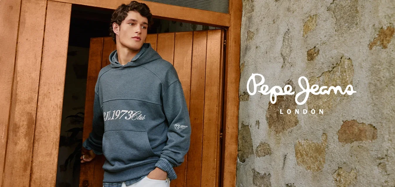 Jonge man in een grijze hoodie en witte broek die een houten deur opent naast een getextureerde stenen muur, terwijl hij opzij kijkt.