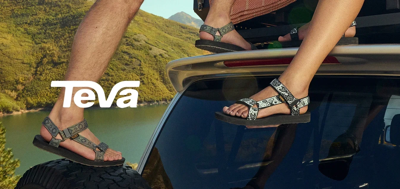 Twee paar sandalen met gevlochten banden, een grijze en een zwarte. Beide hebben een platte zool, die vastgemaakt is met verstelbare gespen, op het dak van een auto.