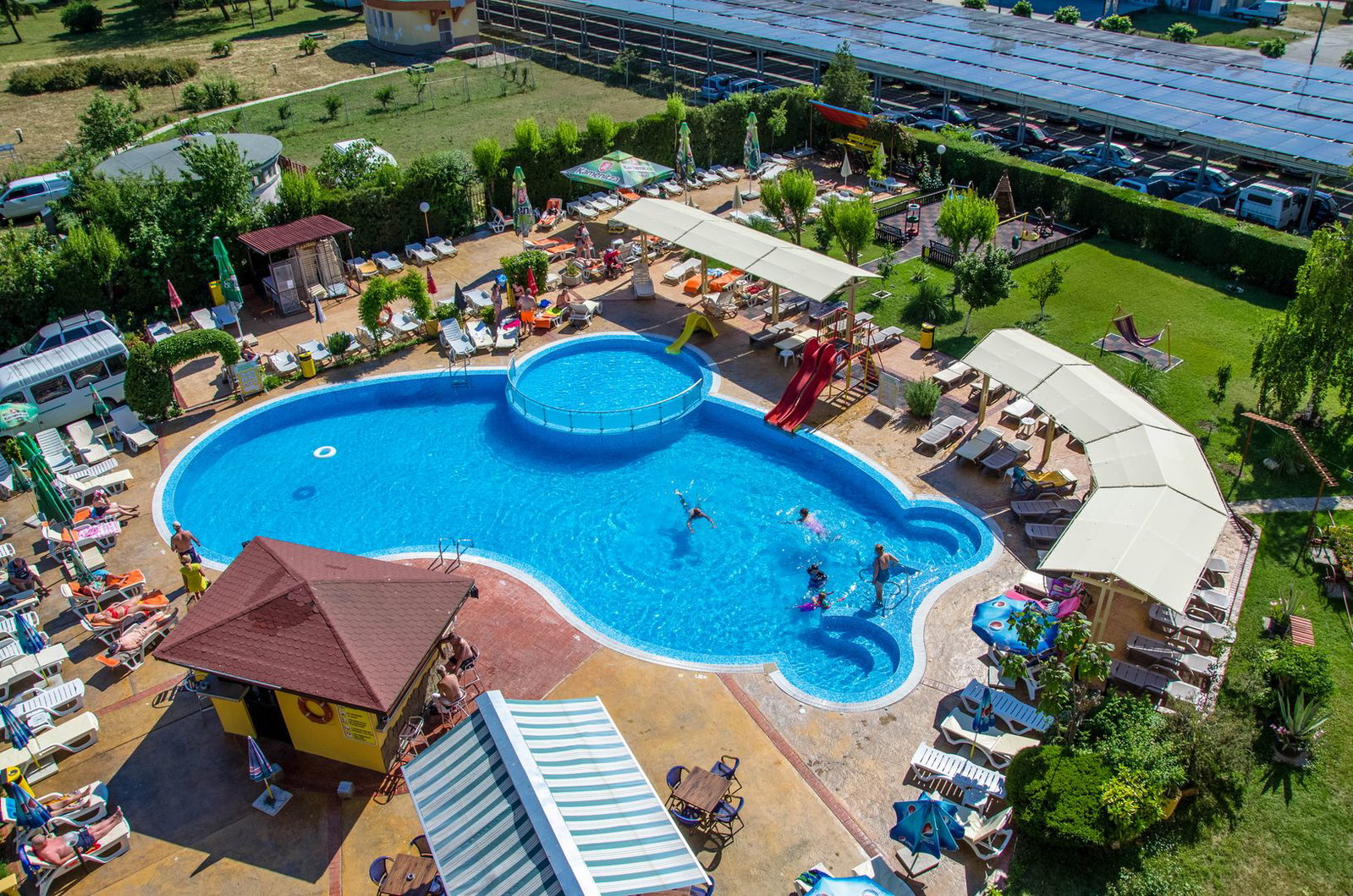 Aerial view of a sunlit outdoor resort pool with children on slides, adults swimming, sunloungers and shaded pergolas around