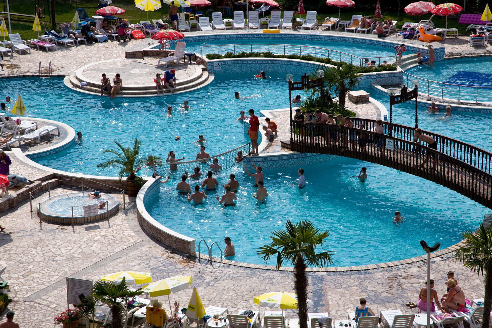 Outdoor resort with interconnected turquoise swimming pools, wooden footbridge, sunloungers, umbrellas and people enjoying swimming