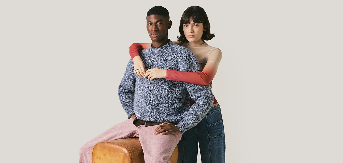 Ein Mann in einem blauen Pullover und pinken Hosen sitzt auf einem Sprossenreck, während eine Frau in einem roten Oberteil und weiten Jeans hinter ihm steht und ihn umarmt.