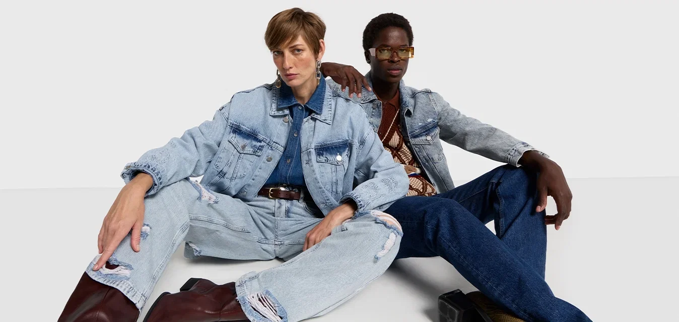 Zwei Models sitzen auf dem Boden, tragen Denimjacken und zerrissene Jeans. Das erste Model hat braune Haare, das zweite trägt eine Brille und ein gemustertes Oberteil.