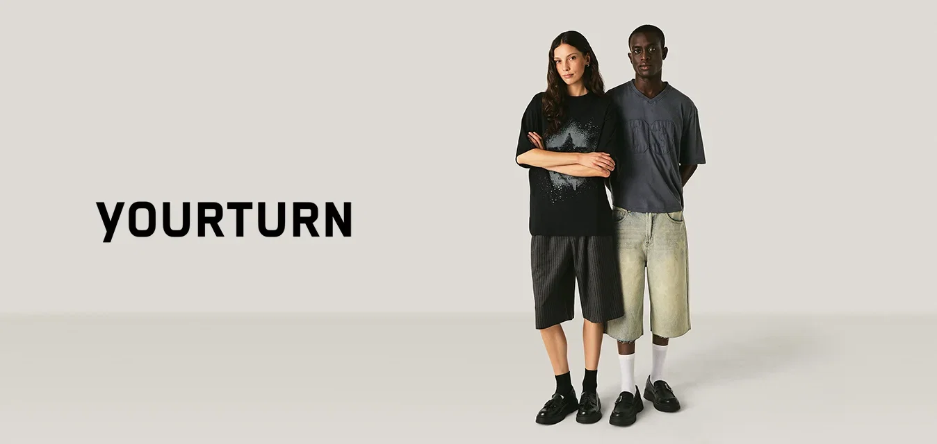 Twee mensen staan zij aan zij, gekleed in oversized t-shirts en knielange shorts tegen een effen lichte achtergrond.