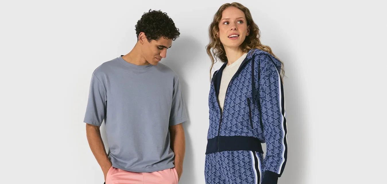 Mann in grauem T-Shirt und rosa Hose steht neben Frau in blauer, gemusterter Zip-up-Jacke und passender Hose mit weißen Streifen.