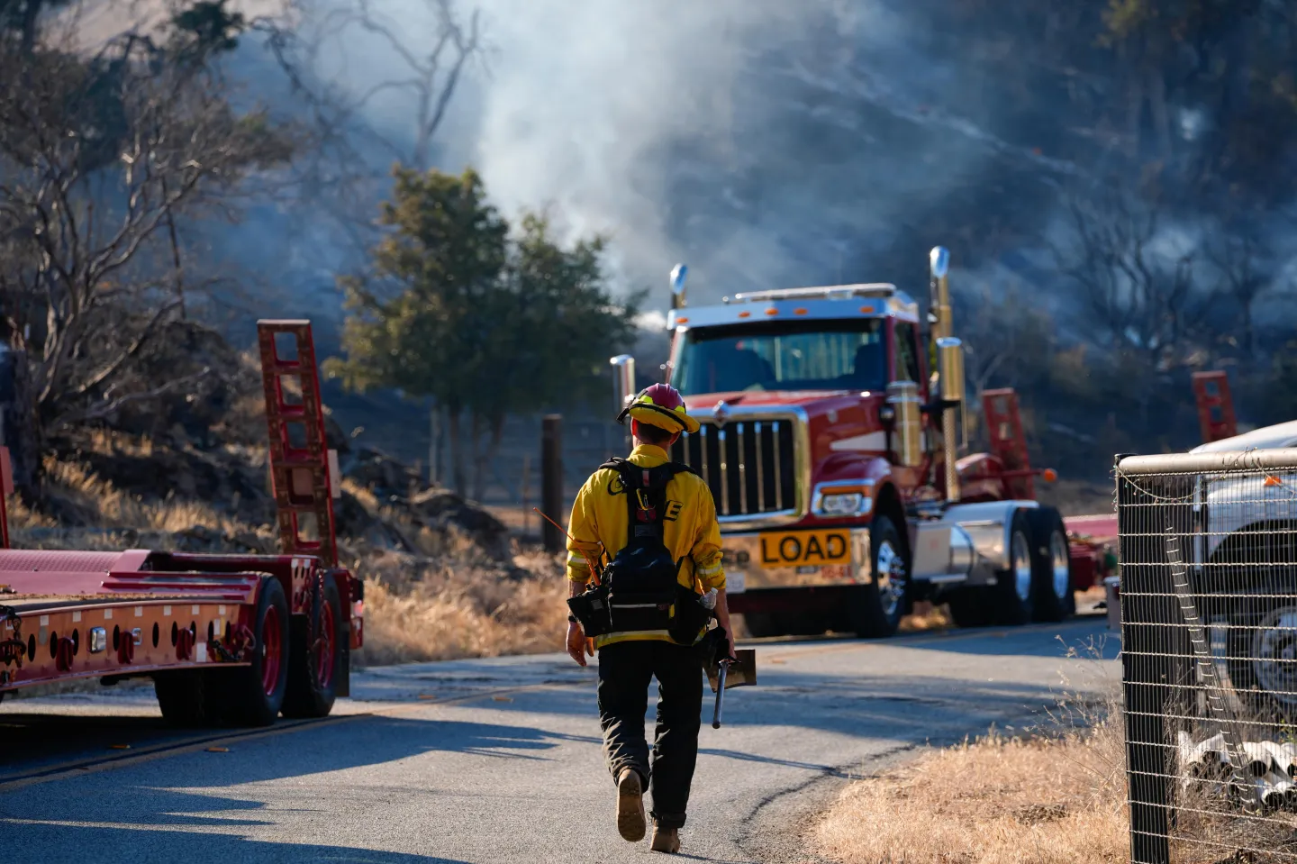 A firefighter wearing a yellow shirt and a red helmet walks down a a road in Santa Clara County, California, toward a red semi truck.