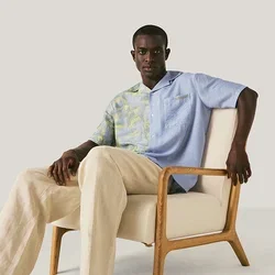 Man in een blauw, patroonhemd en beige broek zittend op een lichtgekleurde gestoffeerde stoel tegen een effen achtergrond.