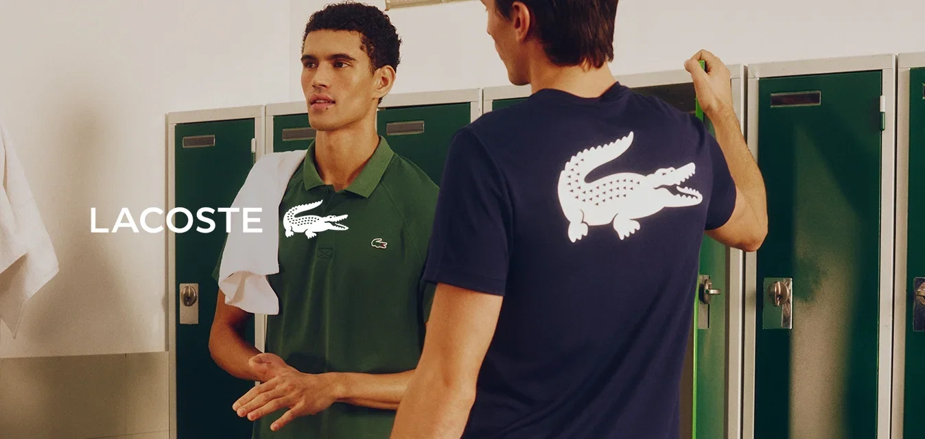 Twee jonge mannen praten in een kleedkamer, één draagt een groen shirt en houdt een witte handdoek vast, de ander een marineblauw shirt met een groot wit alligatorlogo.