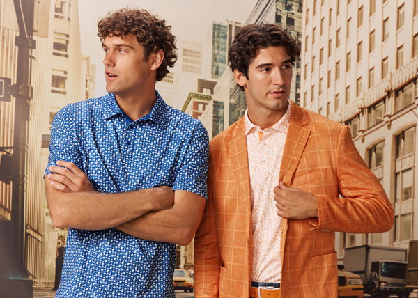 Two models styled for game day, one wearing a blue football-print polo and the other an orange windowpane blazer over a patterned polo, against a city backdrop.