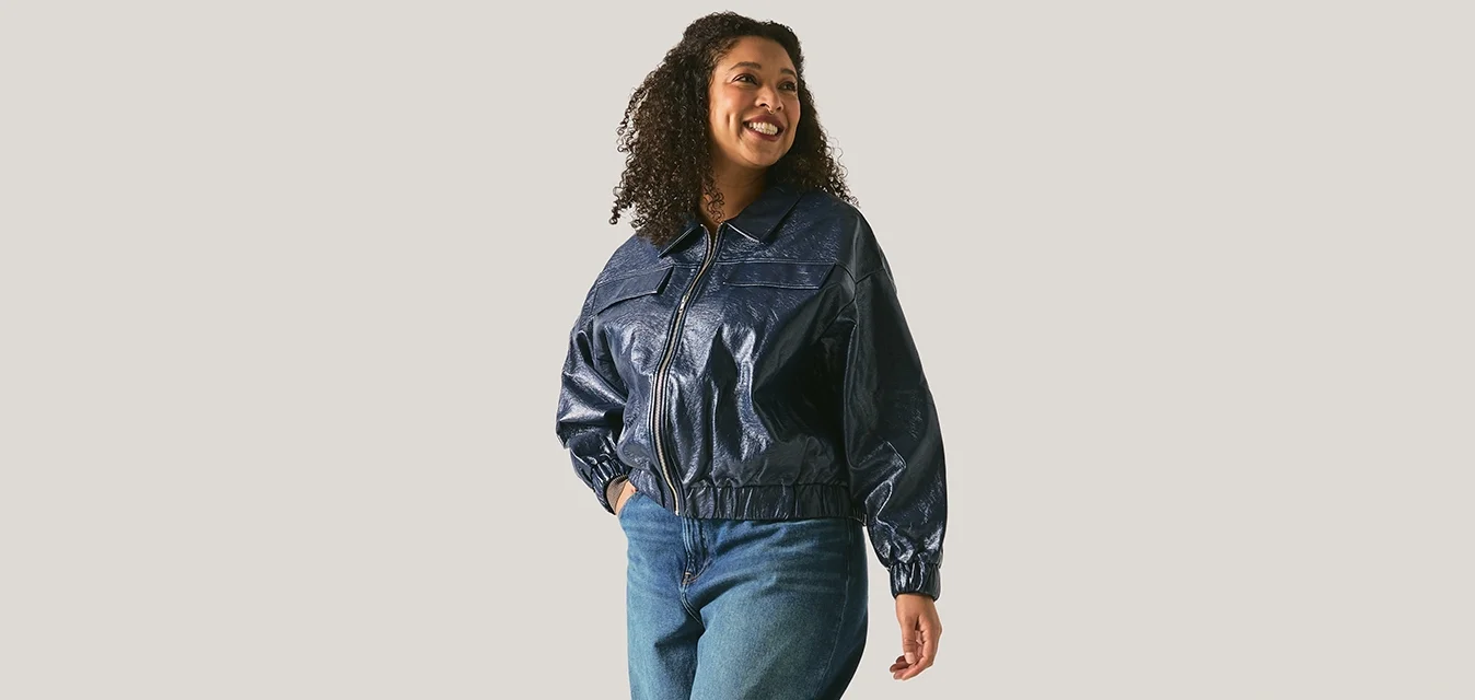 Frau mit lockigem Haar, die eine dunkle Jacke, blaue Jeans mit weitem Bein und schwarze Stiefel trägt, geht lächelnd vor einem einfachen hellen Hintergrund.