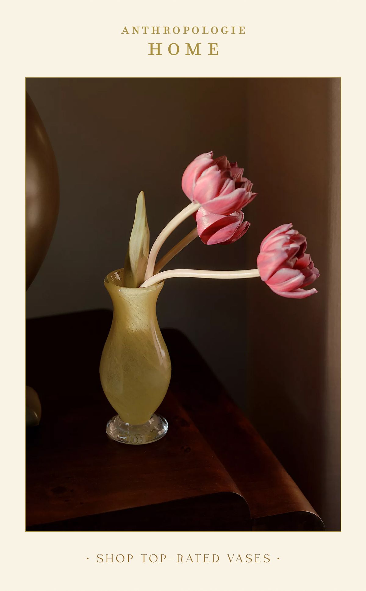 Amber-yellow swirled glass vase holding three pink tulips sitting on a dark wooden tabletop. Text: ANTHROPOLOGIE HOME — SHOP TOP-RATED VASES.