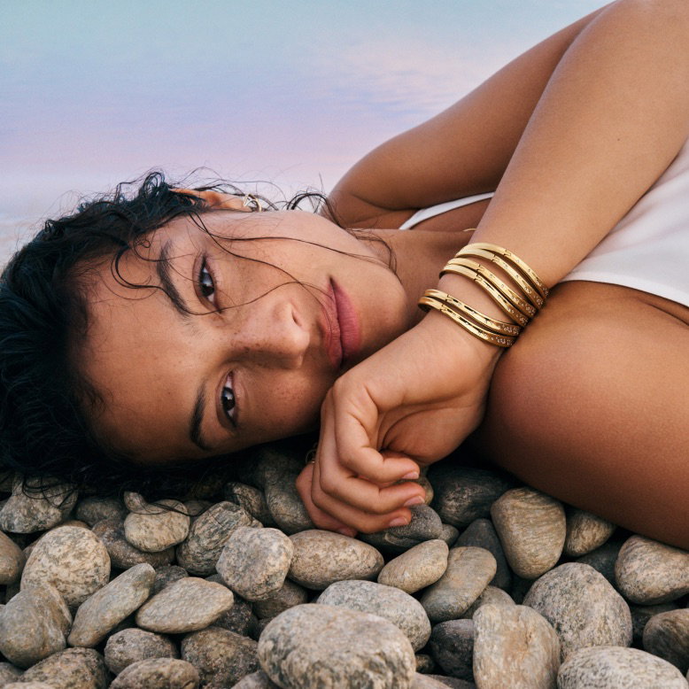 A model rests on pebbles wearing stacked 14k gold-plated bangles.