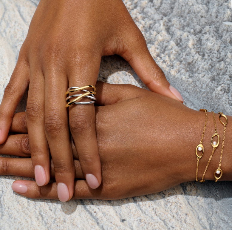 A pair of hands overlap and display an intertwined ring and pearl bracelet.