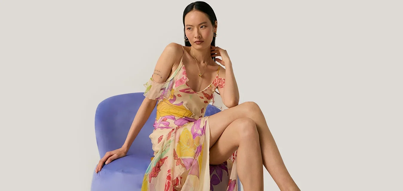Frau mit einem transparenten Blumenkleid und roten High-Heel-Sandalen, die auf einem blauen modernen Stuhl vor einem einfarbigen Hintergrund sitzt.