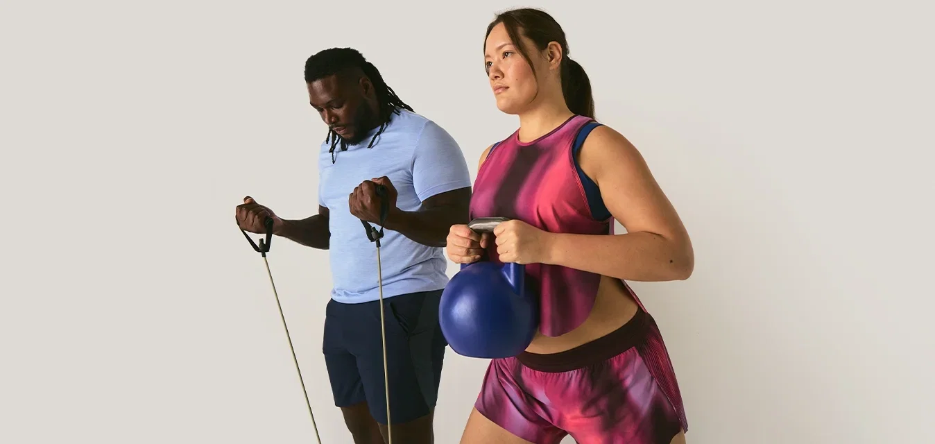 Ein Mann trainiert mit Widerstandsbändern, während eine Frau einen blauen Kettlebell hält und rosa Sportkleidung trägt, vor einem schlichten Hintergrund.