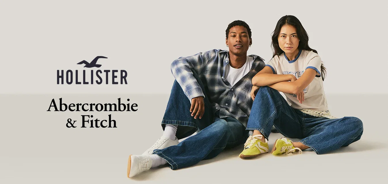 Zwei junge Erwachsene sitzen auf dem Boden, tragen legere Jeans und Turnschuhe und schauen mit entspannten Gesichtsausdrücken nach vorne vor einem einfarbigen Hintergrund.