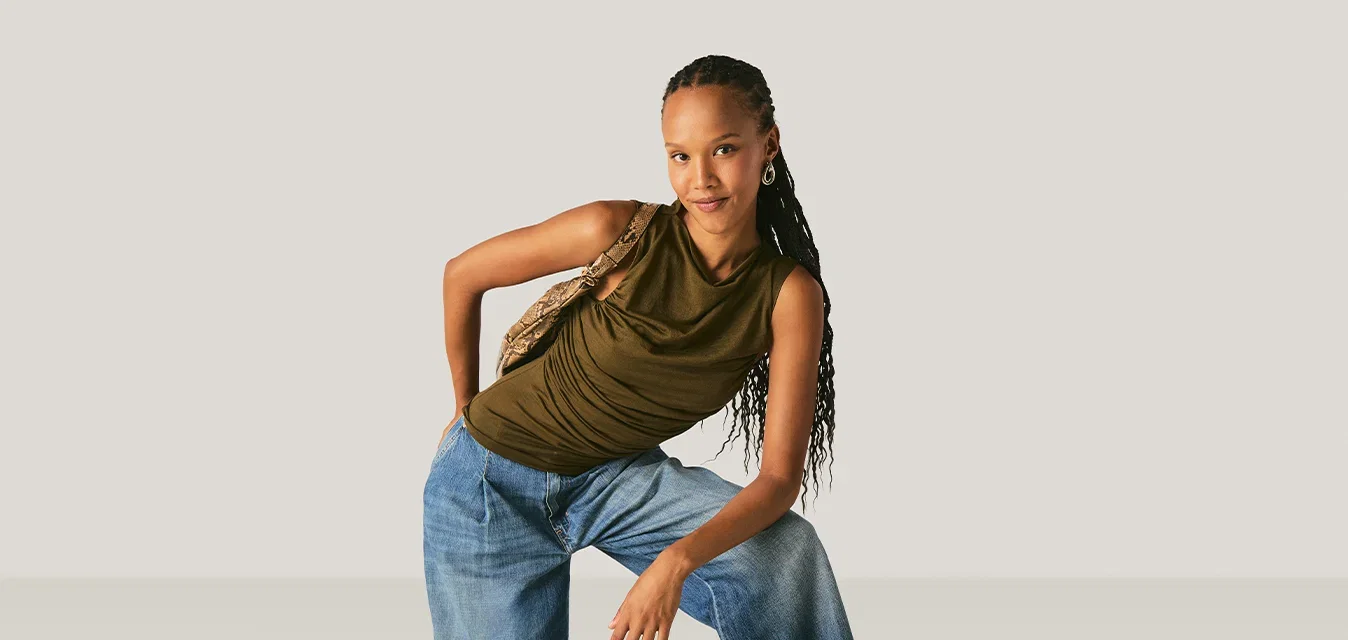 Junge Frau mit langem geflochtenem Haar, die ein ärmelloses olivgrünes Top und weite blaue Jeans trägt und mit einer gemusterten Schultertasche posiert.