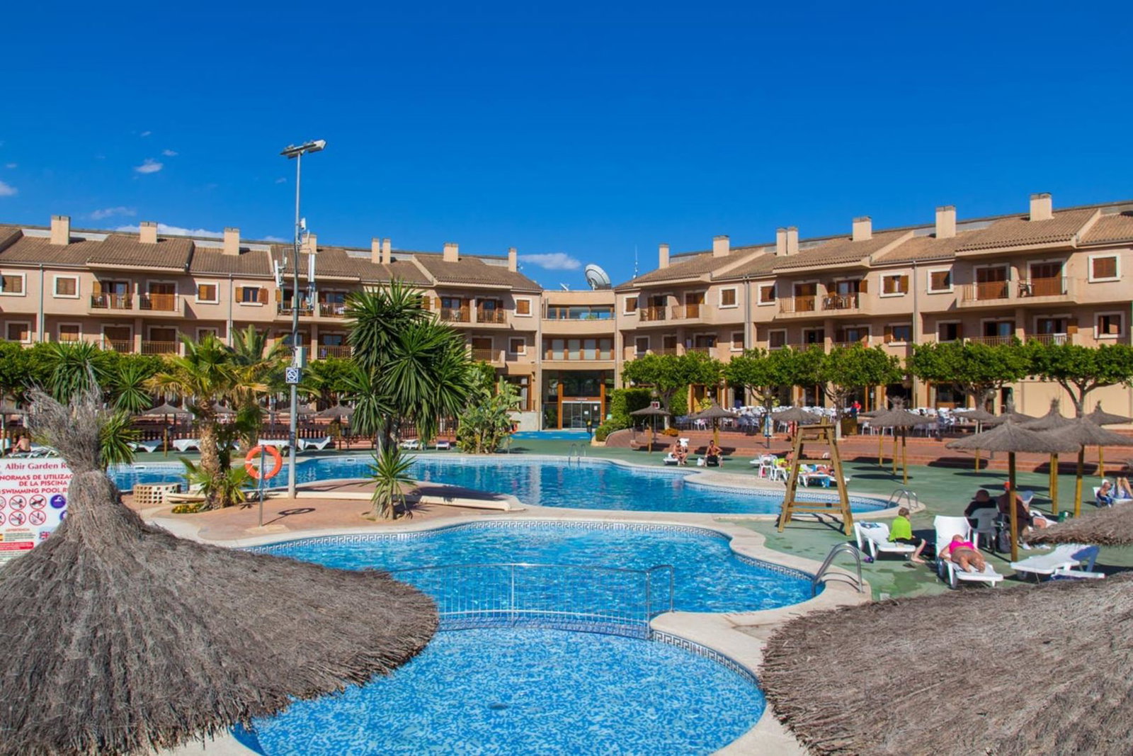 Outdoor resort swimming pools with sun loungers and straw parasols in front of terraced holiday apartments
