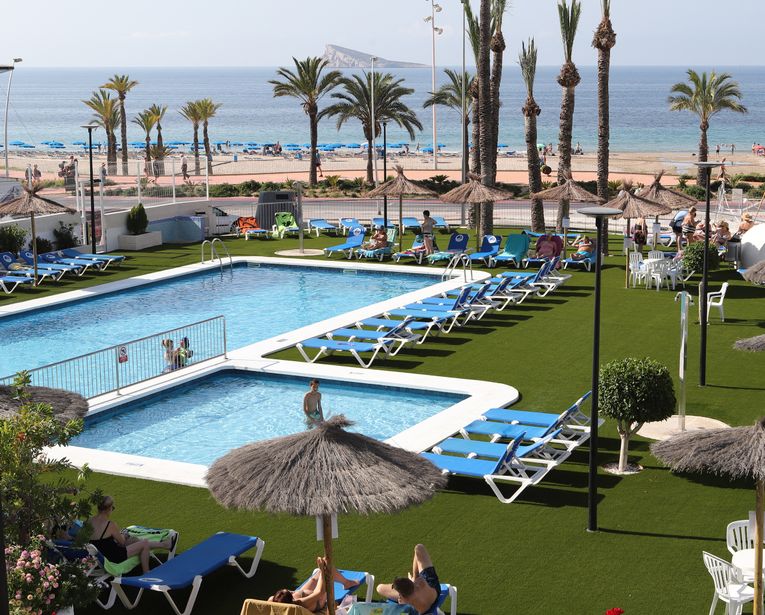 Resort pools with blue sunbeds on artificial grass, thatched parasols and palm trees, sandy beach and sea beyond